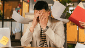 A stressed man at a desk has papers playfully placed on his head, with additional paperwork scattered around him.